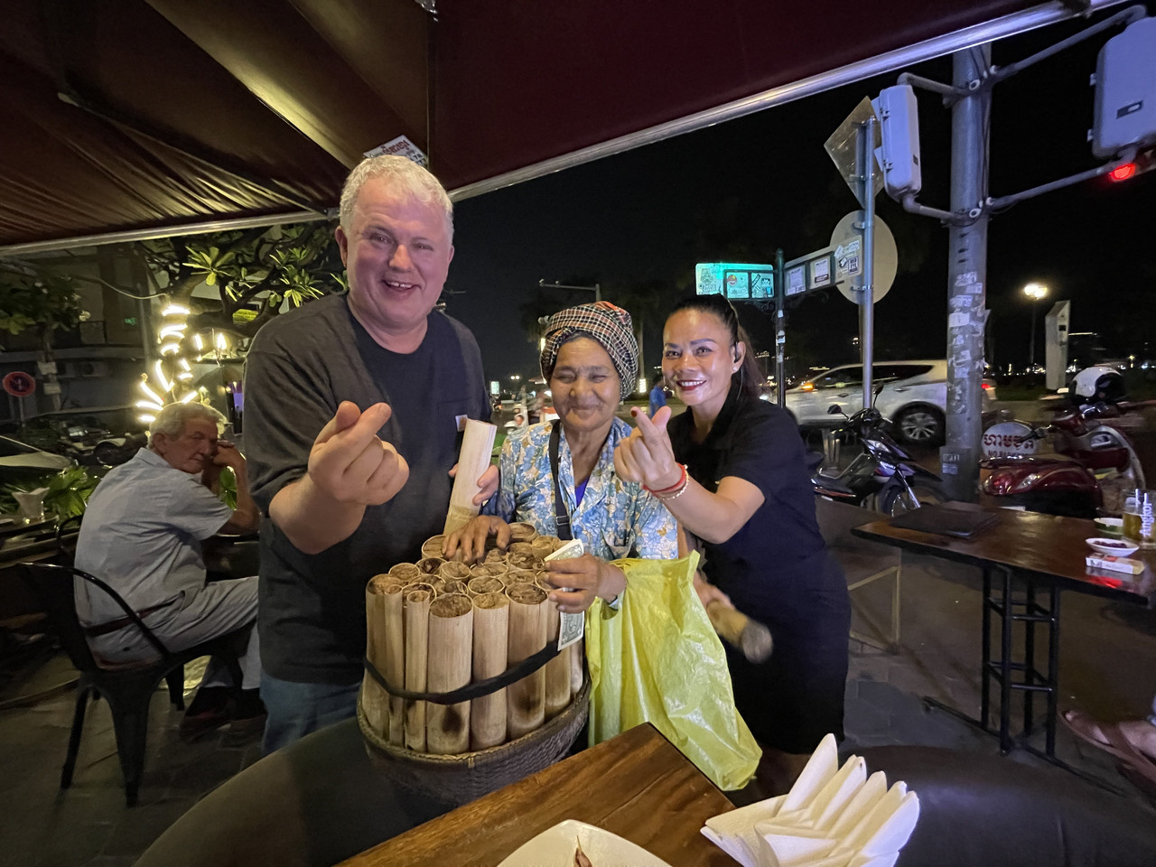 Dr. Phillip Bailey smiling outdoors at night while standing next to a street vendor and another woman around a wooden basket filled with bundled bamboo cylinders. Dr. Bailey and the woman to the right are both making hand-heart gestures toward the camera