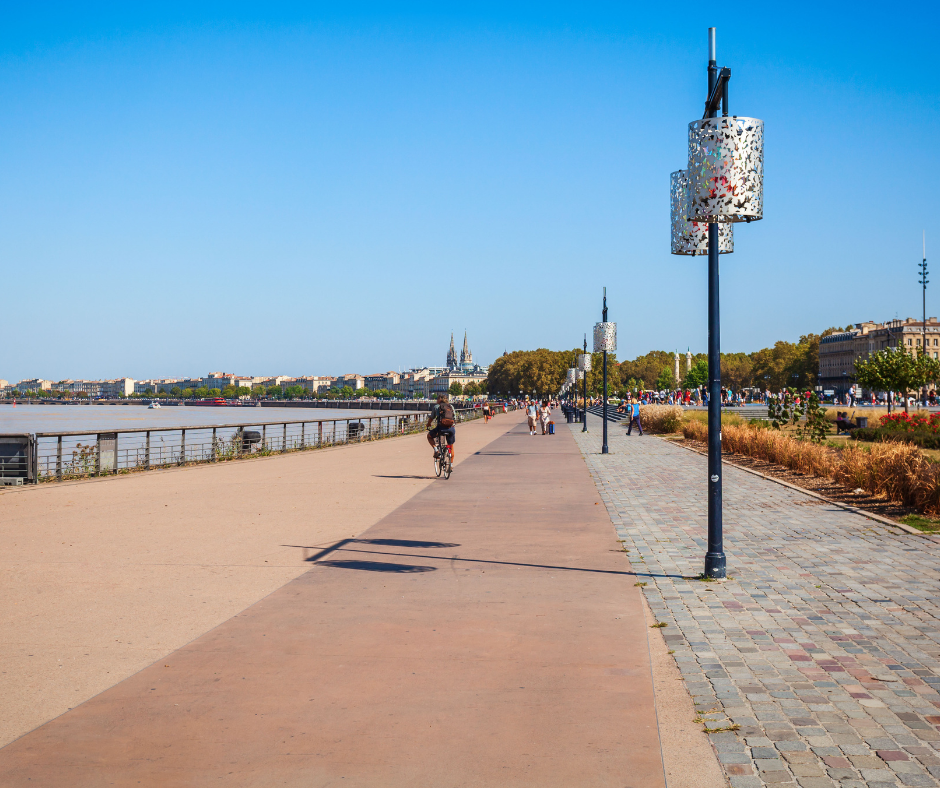 Scenic riverside promenade in Bordeaux, France, with people walking, cycling, and enjoying a sunny day, featuring modern lampposts, a clear blue sky, and historic buildings in the background.