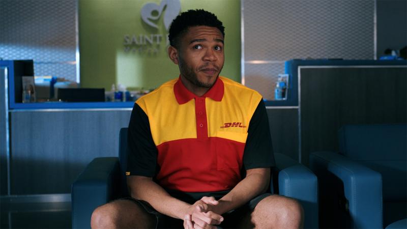 still frame from pluribus showing an unnamed guy sitting down in the otherwise empty lobby of a hospital. He is wearing a DHL delivery uniform but he is acting as a speaker for the collective hivemind here