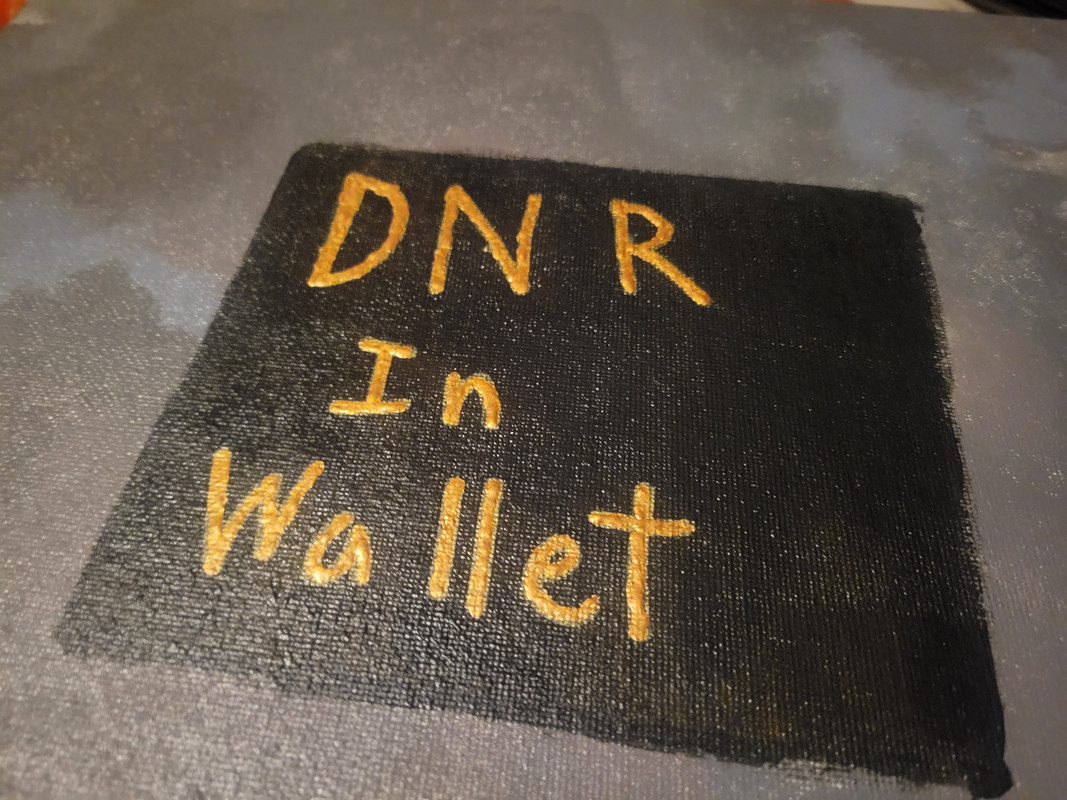 A close up image of an acrylic painting of a black square on a gray background. The black square takes up most of the picture and has gold text on it that read DNR In Wallet. The gray background is made of many various shades of gray in rough blob like shapes