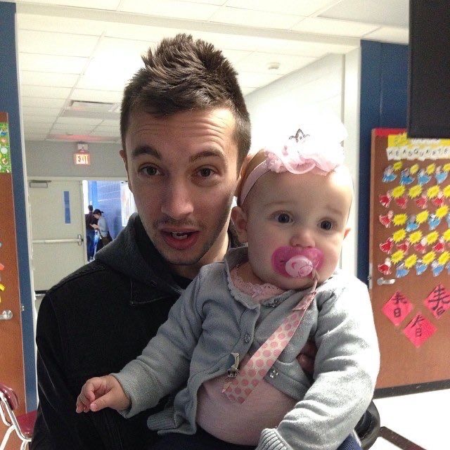 [A young Tyler is holding a baby girl to his chest with both of them facing the camera. It looks like he was caught off guard that the photo was being taken, his eyebrows are raised up and his mouth is slightly open like he was going to say something. There are stood in a hallway of a building/church