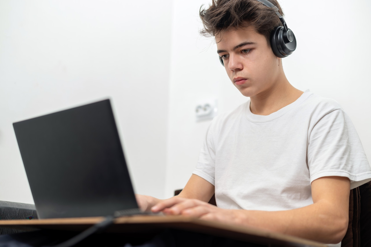 el muchacho adolescente esta usando la computadora portatil con los auriculares en casa rostro serio