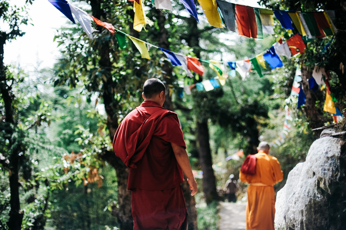 Twee monniken lopen in Nepal over een pad een berg af, langs natuurlijke rotsstukken. Boven hen hangen de typische slingers van gekleurde lappen stof.