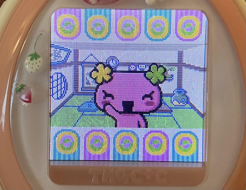 close up photograph of a tamagotchi plus color device. furawatchi has come close to the camera and is smiling and waving at the viewer
