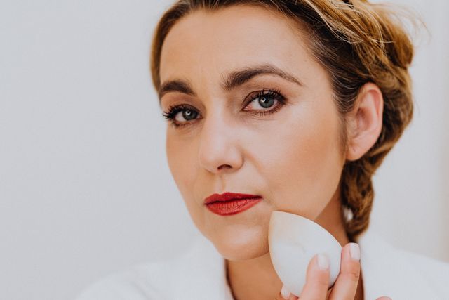 a woman with cakey, patchy foundation on her face holding beauty blender to blend the makeup 