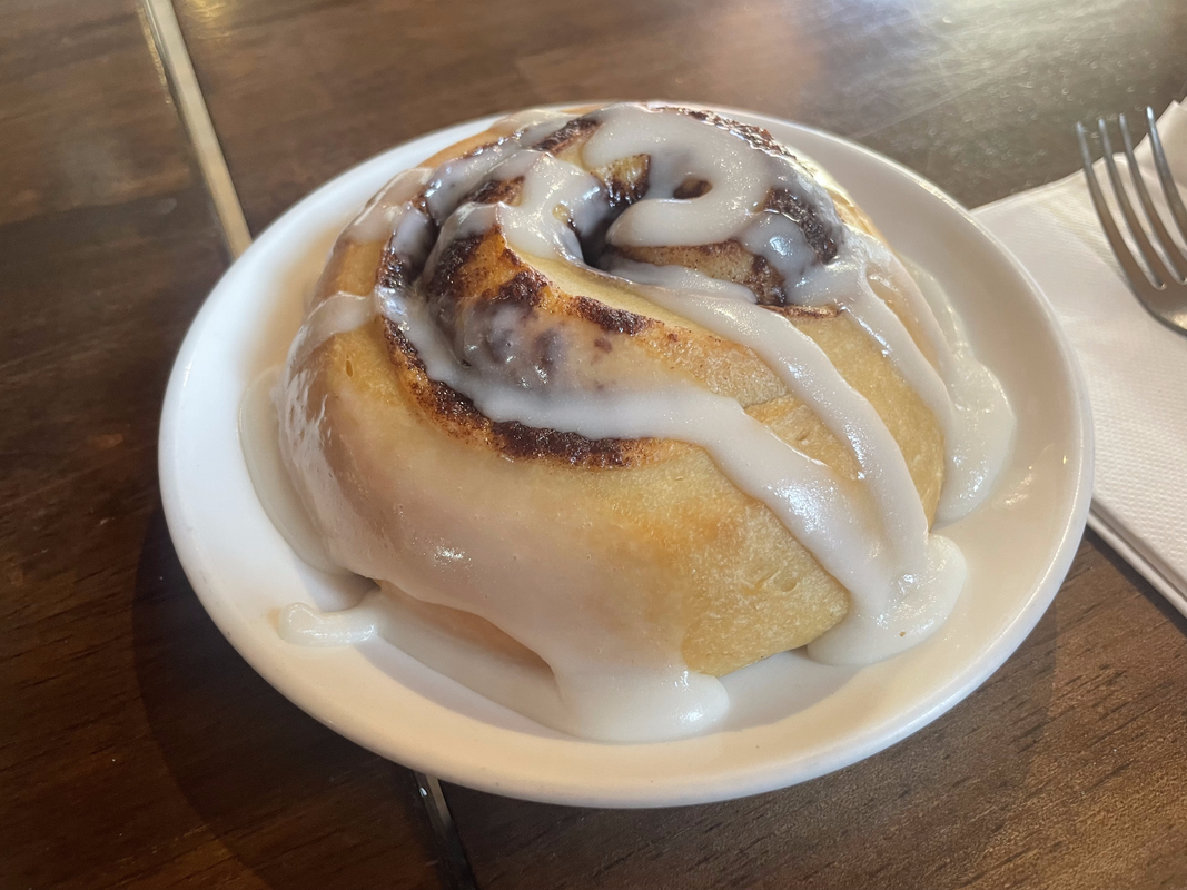 a photo of a glazed cinnamon roll on a white plate on a dark wooden table!
