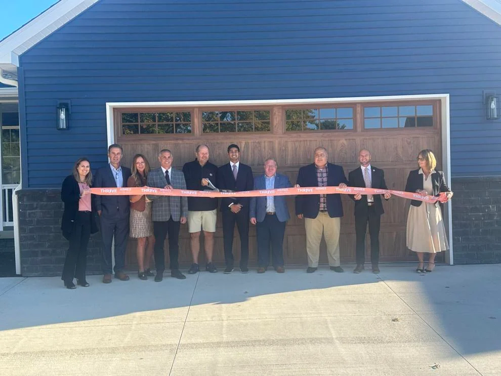 A group of people pose holding a long red ribbon and pair of scissors.