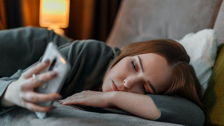 Jovem mulher no sofá focada na tela do smartphone lendo resenhas e indicações no BookTok em um momento de relaxamento.