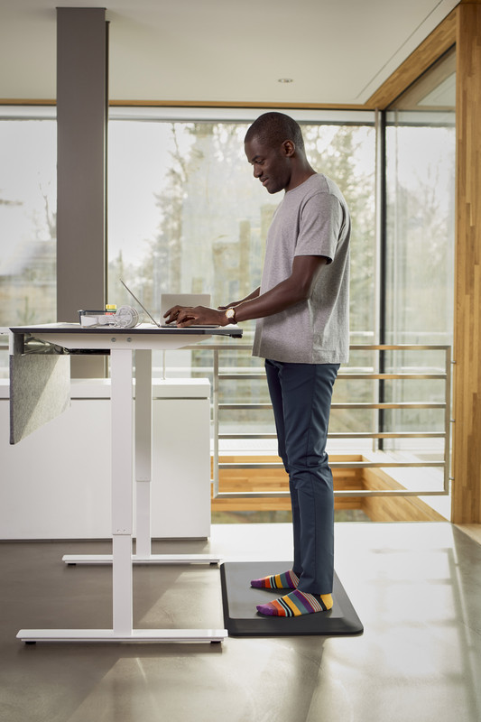 Mann steht auf einer Aktiv-Bodenmatte mit Waldboden-Effekt vor einem Stehschreibtisch.