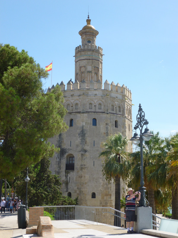 The Torro del Oro is a twelve sided tower, made of what looks like pale stone. There are trees around it, and a Spanish flag flapping in the breeze on top.