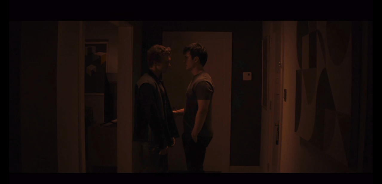 shane and ilya stand in the entranceway of shane's hotel room. the lighting is dark orange with many shadows. ilya is wearing a patchwork black and gray coat and gray pants. shane is in a short-sleeved gray shirt and black pants. 