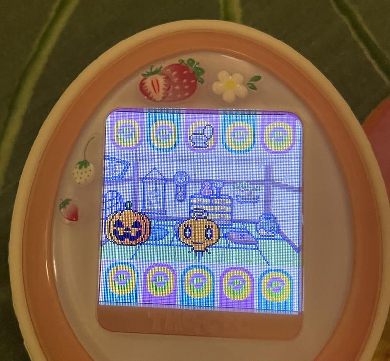 close up photograph of a tamagotchi plus color device. memetchi is standing in her living room next to a pumpkin