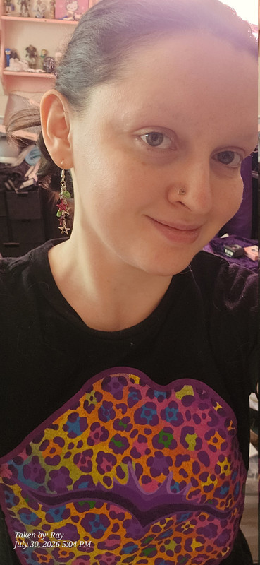 me wearing a black lisa frank shirt. it has a giant print of rainbow lips with rainbow cheeta print. Everything on the print has a purple outline and parts of the purple outline are a lighter purple. my heart shaped trichrome nostril piercing on my left side is visible and I'm wearing dangly star earrings, some are neon green star shaped beads, some silver hollow star shapes and flat textured bright fuchsia with tactile black cheeta print on them.