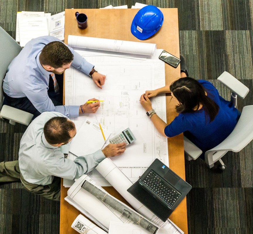 overhead image of 3 people reviewing engineering drawings at a table and doing calculations