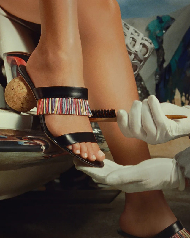 Ferragamo Pre-Fall 2025: Un Tocco di Colore Artigianale ai Piedi Un piede indossa un sandalo Ferragamo mules nero della collezione Pre-Fall 2025 con tacco e una fascia frontale decorata con frange multicolore vivaci. Mani guantate di bianco puliscono o sistemano la scarpa.