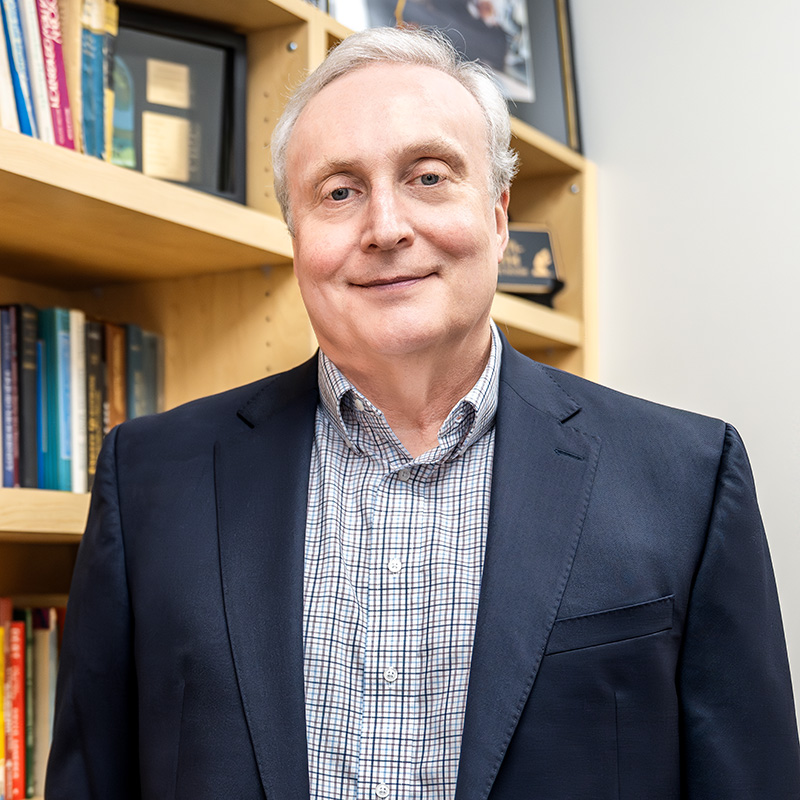 Headshot of Doug Corbitt smiling in a navy blazer and plaid button-down shirt in front of a wooden bookshelf.