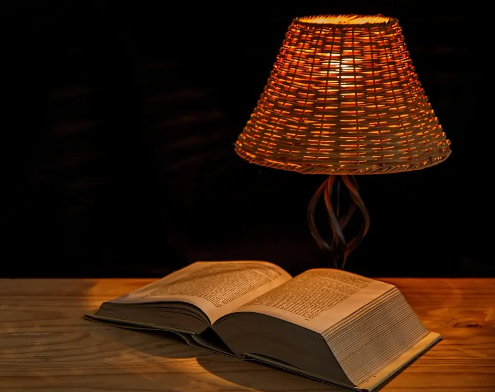 Lampe de chevet en rotin allumée à côté d’un livre ouvert sur une table en bois, dans une ambiance chaleureuse.
