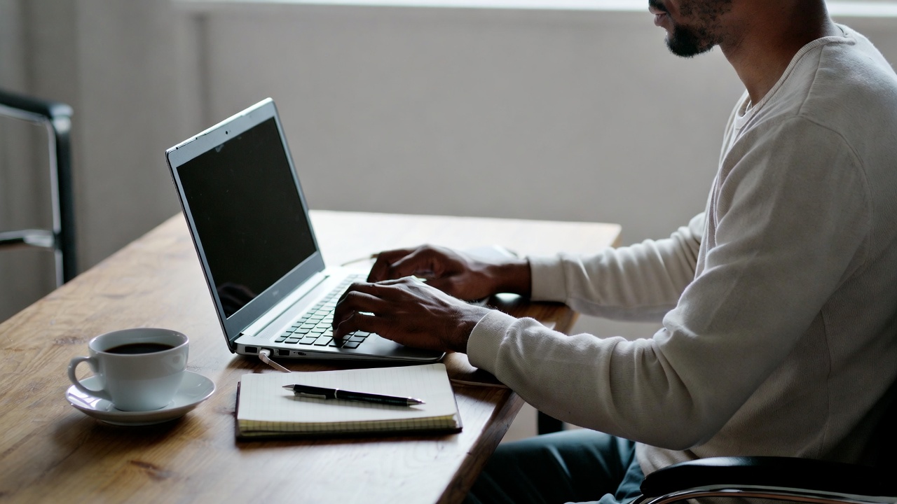 Person working on a laptop at a home office desk with a notebook and coffee