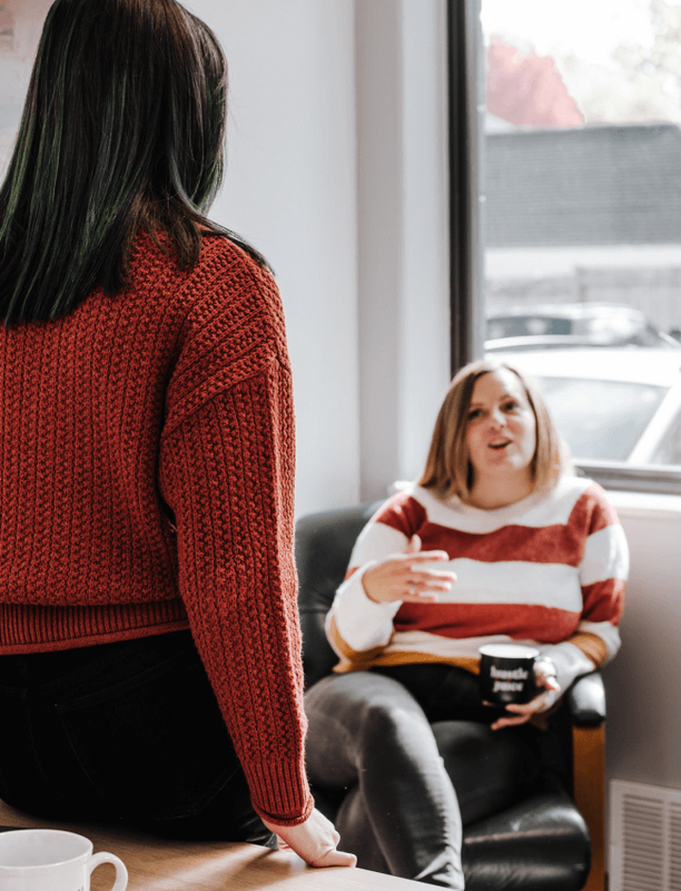 Two women collaborating in an office, one standing with back to viewer, one seated.