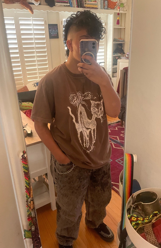 a warm-toned photo taken in a full length mirror of a person in acid-washed jeans and a brown Sap band t-shirt, with a wallet chain reaching from hir front belt loop to hir back pocket. zie have a newly cut mohawk haircut-- shaved really close on the sides and big and fluffy along the center