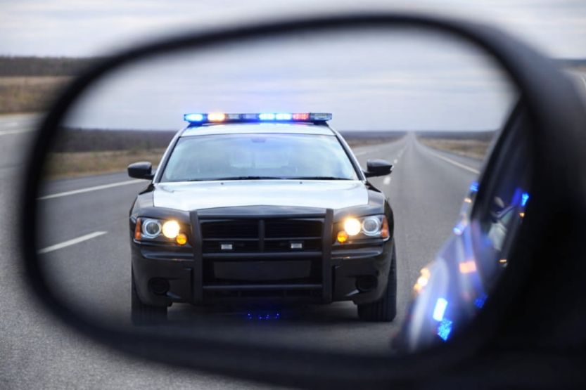 a patrol car approaching with lights on, reflected in a vehicle’s side mirror