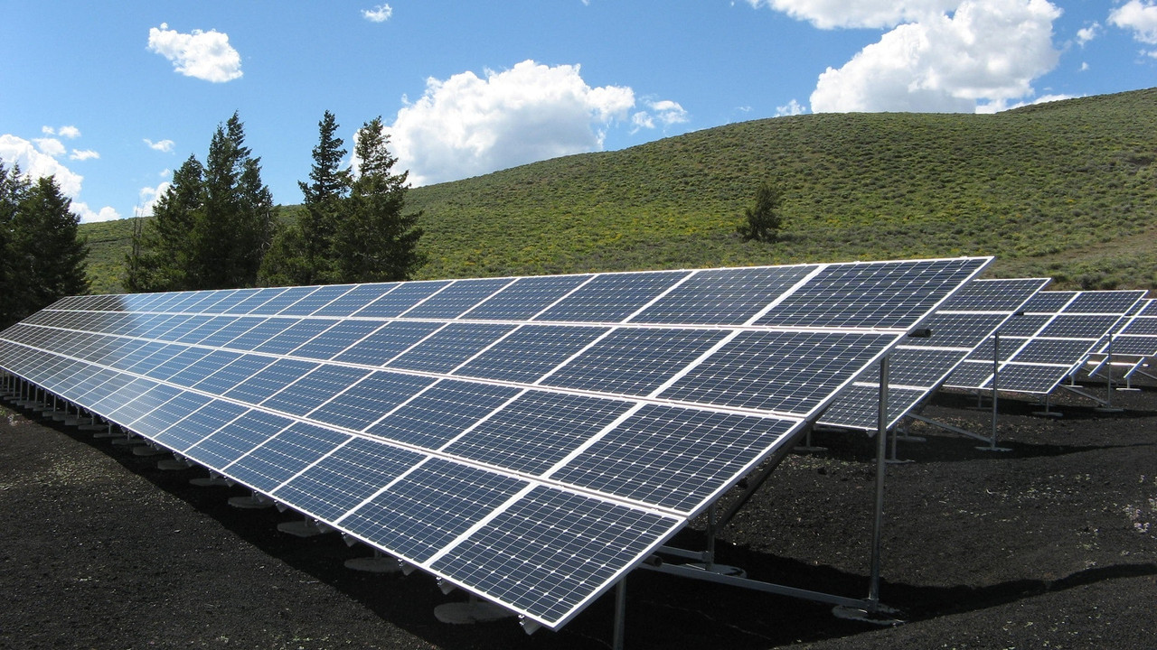 row of solar panels at an angle with green hill and bright, slightly cloudy sky in the background