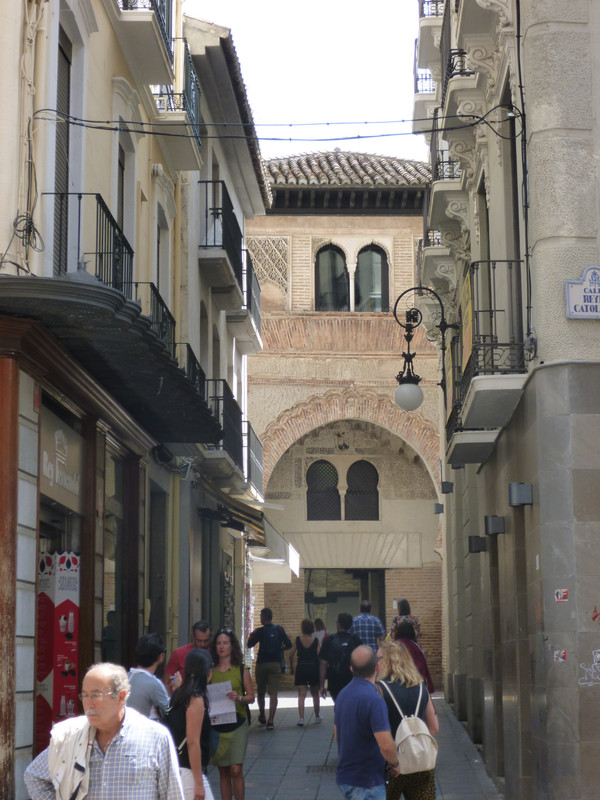 Left and right are rows of shops.  In the front of the photo are people walking along the street.  At the back is an orange stone arch in mudejar style.