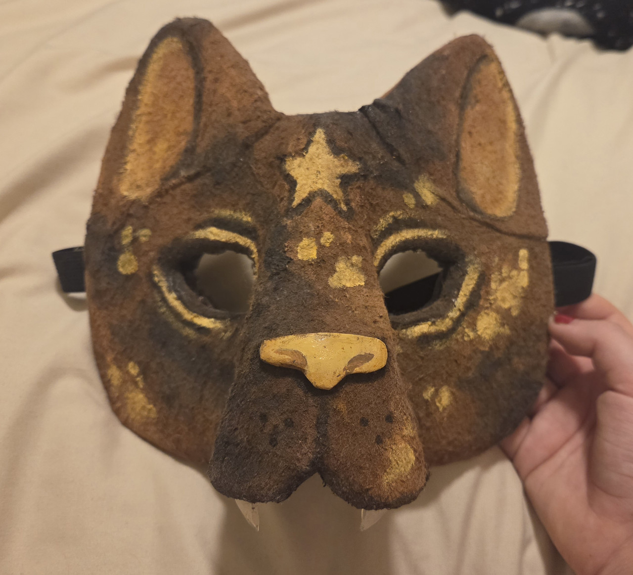 1 of 3 pictures of a cat therian mask. The mask is felted, and painted to resemble a tortoiseshell patterned cat, with extra bright yellow splotches. There is a star in the center of its forehead, and its eyes are empty.
