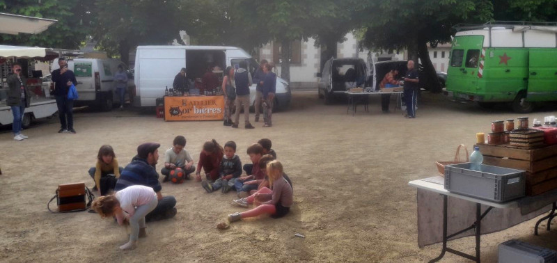 Colin le conteur qui conte assis par terre entouré d'enfants sur un marché de plein air