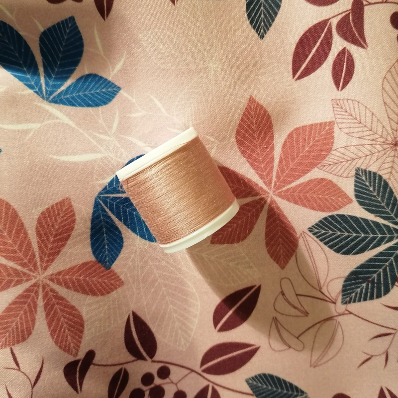 photo of a dusty pink fabric with printed leaves. on it lies roll of thread almost the same color as main shirt.