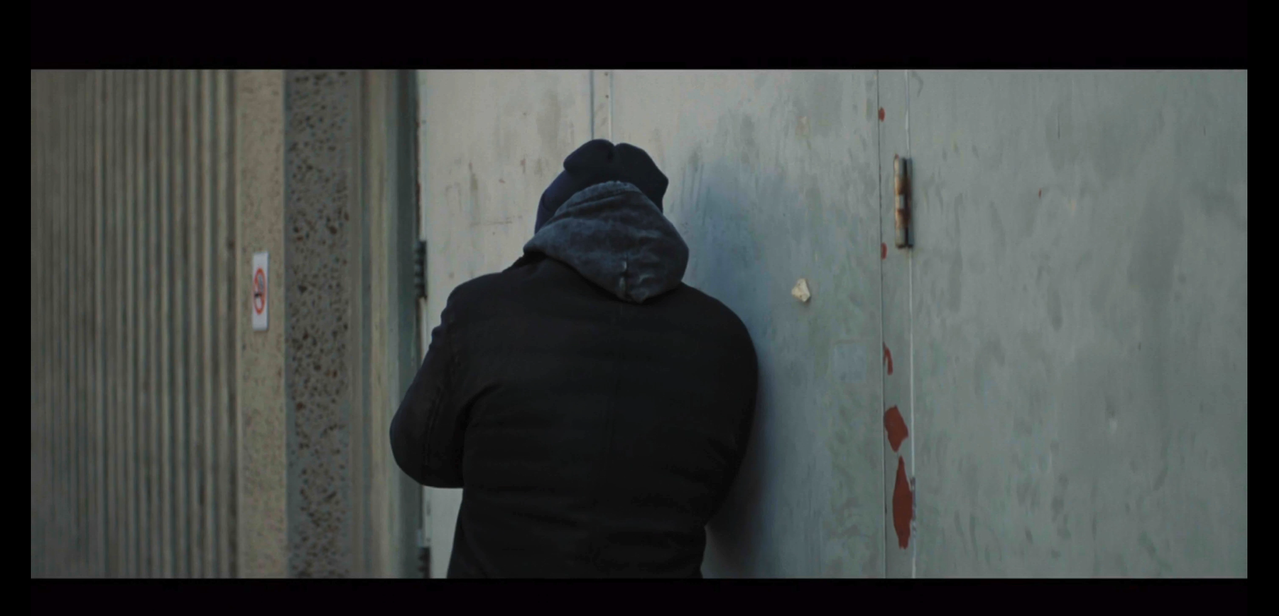ilya stands with his back to the camera, leaning against an exterior wall. he's wearing a black beanie/toque and jacket, with a dark gray sweatshirt hood.