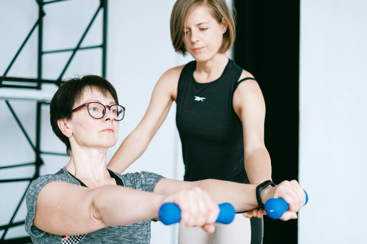 Midlife woman doing a dumbbell deadlift in a gym with coach support