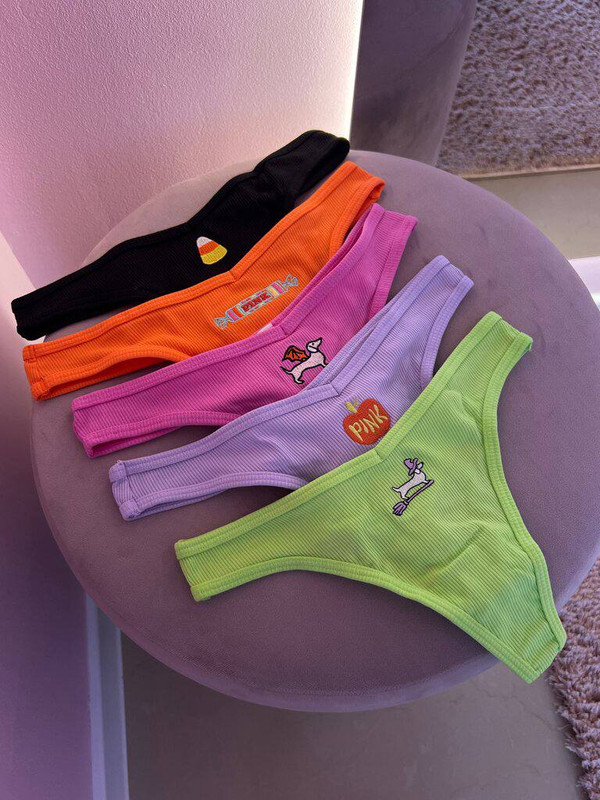 Четыре женских трусика-стринги в рубчик (чёрные, оранжевые, сиреневые и салатовые) разложены на сером пуфе; на каждой модели небольшая вышивка/нашивка.