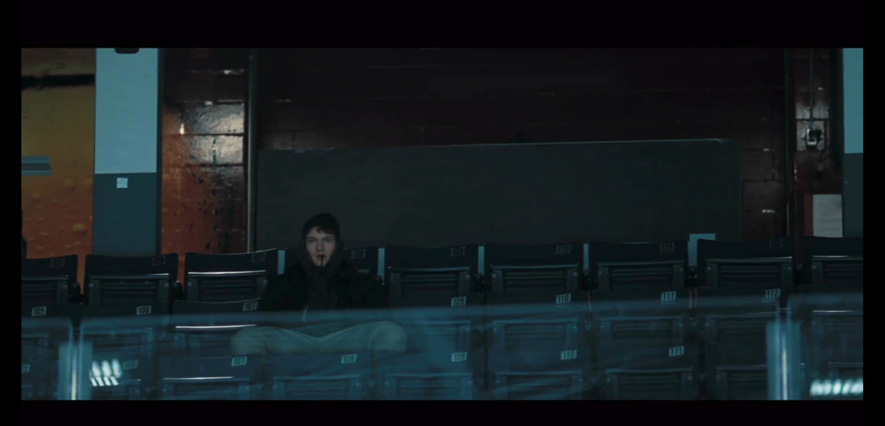 ilya sits alone and off-center in the arena seats. he's chewing on a sweatshirt string with his hands in pockets, knees spread wide, and his hood pulled up over his beanie. he's wearing a black beanie, dark gray sweatshirt, black coat, and medium-gray sweatpants.