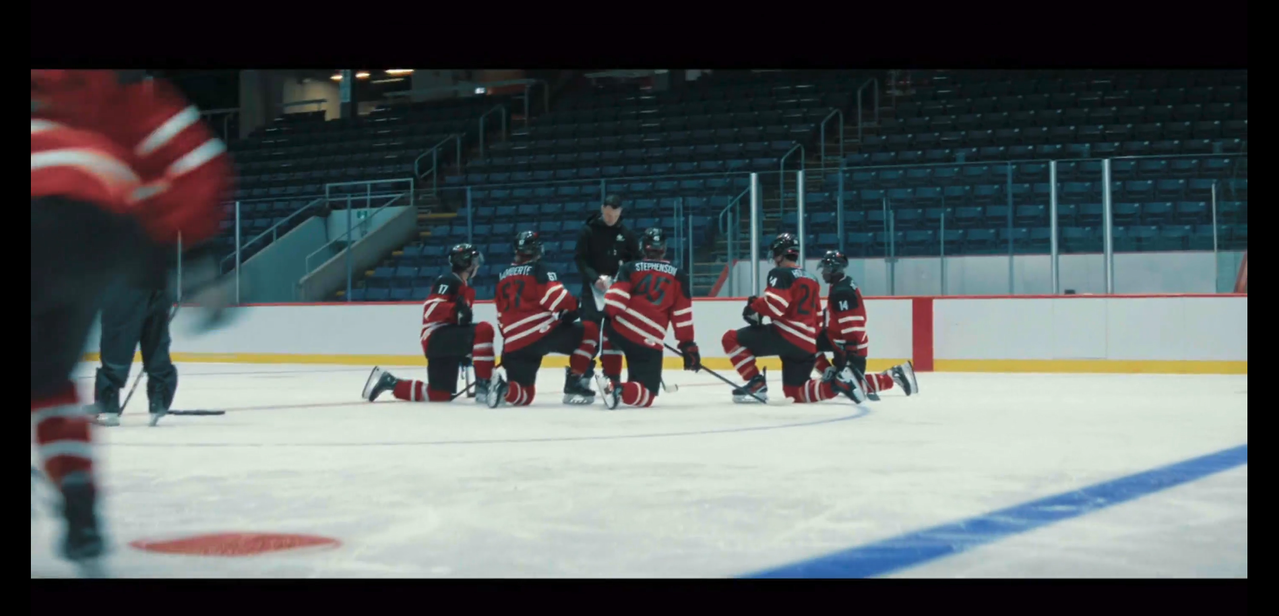 5 players of the canadian youth national hockey team take a knee at center ice in front of their coach. their uniforms are red, white and black. their jerseys have a red base, white stripes at the elbows and waist, white lettering for the names, and black shoulders and lettering for the numbers. their shorts and helmets are black. their socks are red with two white strips mid-shin.