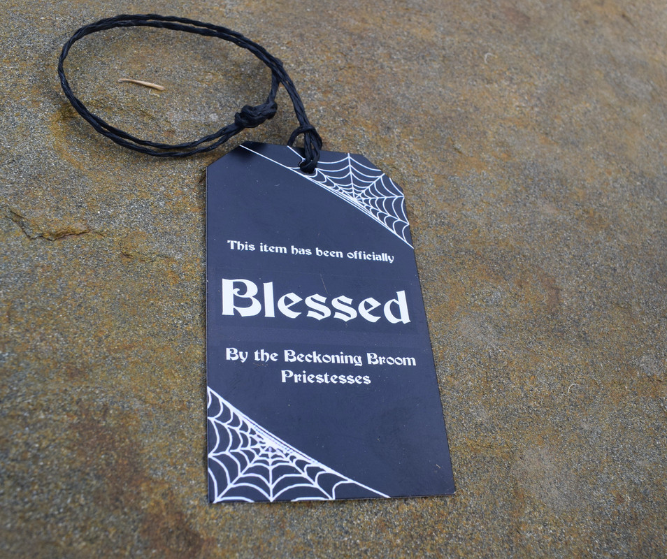 Black tag with spider web pattern saying This item has been officially blessed by the Beckoning Broom priestesses resting on rock