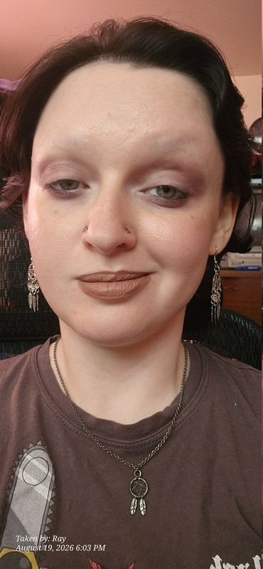A selfie of me wearing a grunge makeup look and smirking with pentagram dream catcher earrings and a tiny pentagram dream catcher necklace my two heart shaped nostril jewelry pieces are also visible and the black butler anime print on my brown tee shirt is almost visible