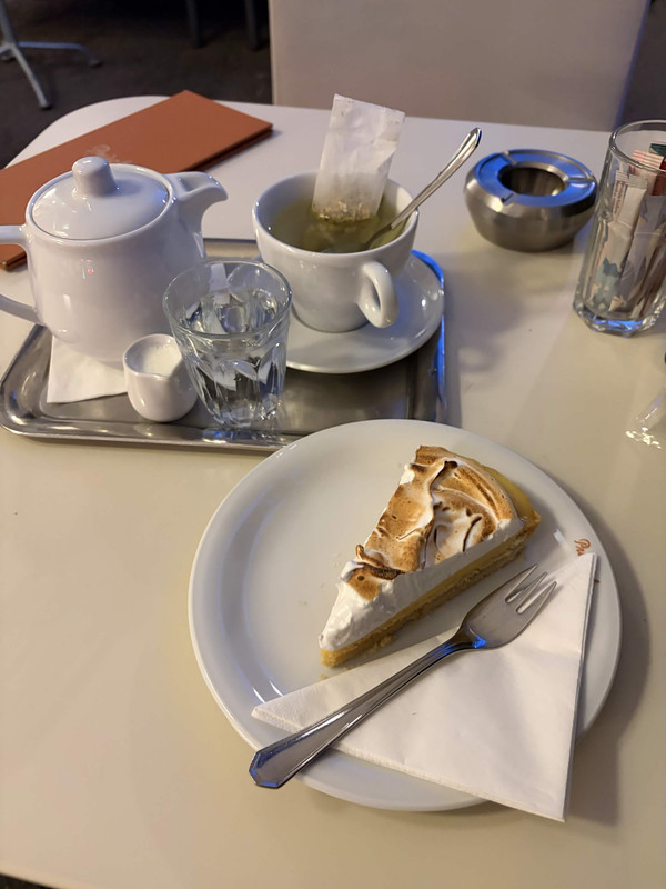 my night time tea in vienna. a metal tray with a small white ceramic tea pot, tea cup, a minature serving cup of milk and a glass of water sit on it. in the foreground is a plate of lemon meringue. to the very side is a metallic ashtray and a glass cup holding paper sugar tubes