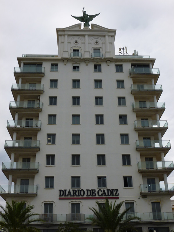 Multi-storey building. White fronted, there are four windows with no frames on each floor, with a balcony on either end of the floor. At the bottom are the tops of palm trees, and top there is a three-columned older style top with bronze winged victory on top. Towards the bottom of the building is a black sign saying Diario de Cadiz with a red line underneath.