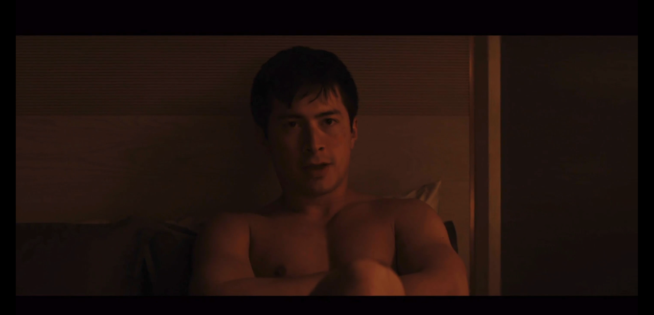 shane sitting up against the headboard, naked.