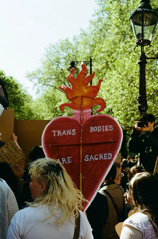 a person holding a burning heart shaped sign that says: trans bodies are sacred.