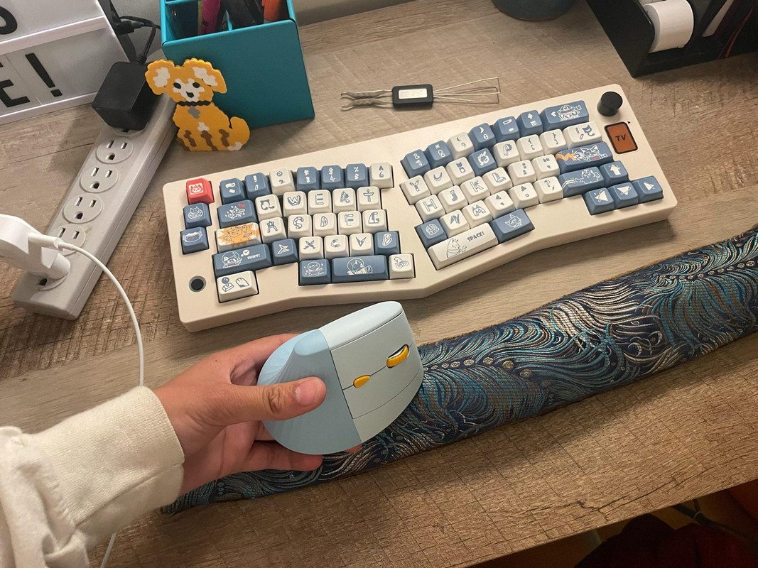 a photo of a desk with a lovely ergonomic keyboard with little cats on the keys and stylized cartoonish letters alongside a light blue ergonomic mouse with a yellow scroller and a little blue/brown bean bag wrist rest thing!