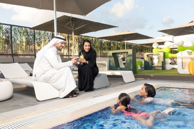 عائلة تستمتع بالسباحة في مسبح Studio M Arabian Plaza Hotel & Hotel Apartments في دبي، حيث يضحك الأطفال بجوار والدتهم ووالدهم في أجواء مرحة.