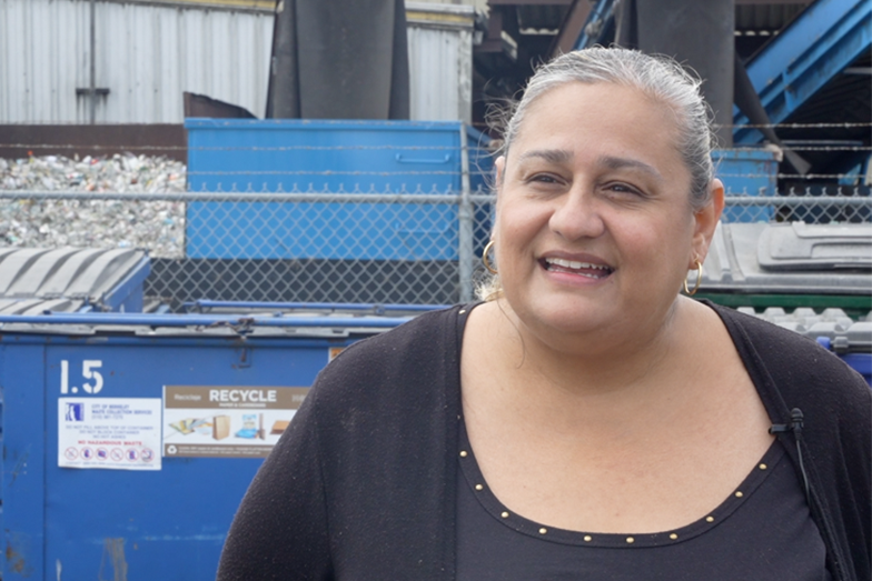 Leticia is smiling and standing in front of waste management equipment.