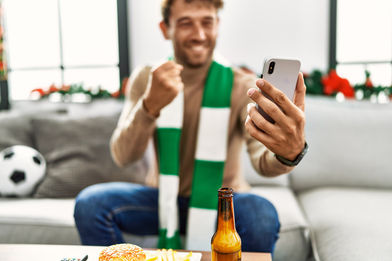 young hispanic man supporting soccer match using smartphone sitting by christmas decor home