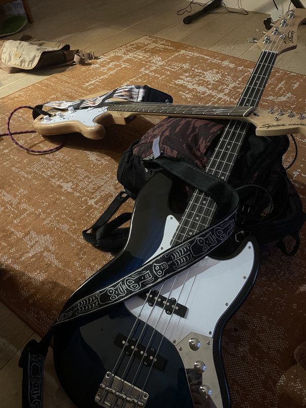 A photo of a blue-black bass guitar with its neck laying against a backpack. A tan wood-stained guitar lays on the backpack with its neck on top of the bass's.