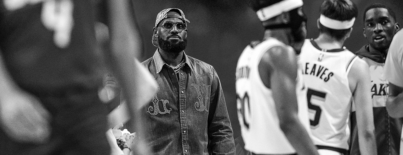 LeBron James, de óculos e jaqueta jeans estilizada, acompanha o jogo da lateral da quadra enquanto observa seus companheiros do Los Angeles Lakers.