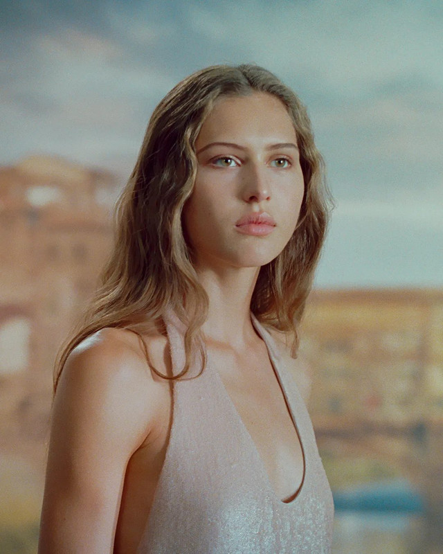 Ferragamo Pre-Fall 2025: Il Volto Raffinato della Nuova Collezione Primo piano di Angelina Kendall nella campagna Ferragamo Pre-Fall 2025 con lunghi capelli ondulati, indossando un top rosa pallido con scollo a V, su uno sfondo sfocato che ricorda un paesaggio urbano.
