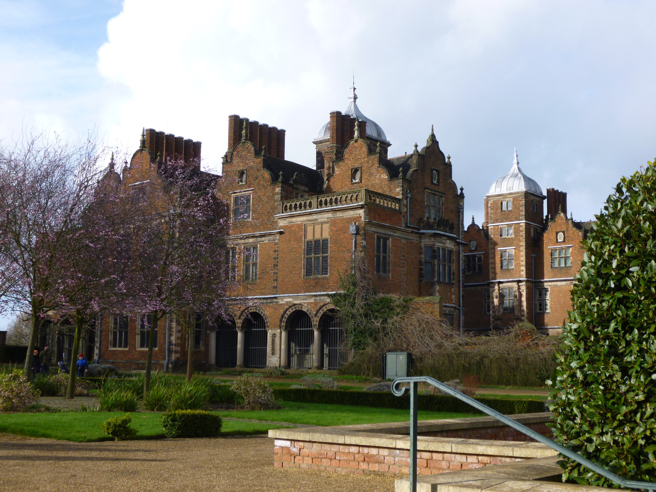 Photo that you can see the front of the hall and the left hand side. From this angle you can see the cupola tops of two of the roof sections which you cannot see in the next photo. The view is from the garden. You can see a hedge in the right hand side of the photo, and trees with purple leaves on the left.
