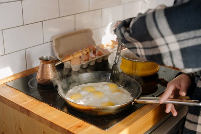 A pan with eggs on a stove along with tea kettle and eggs tray